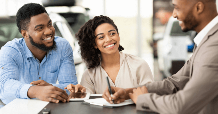 A couple smiling while doing paperwork with a car salesman at a dealership.