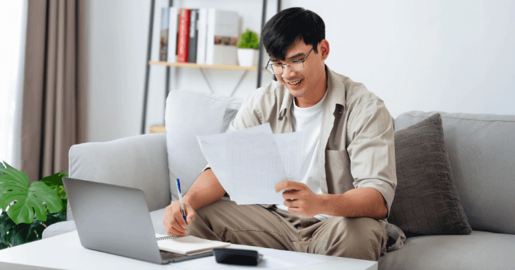 Man sitting on a couch looking a paper with his laptop